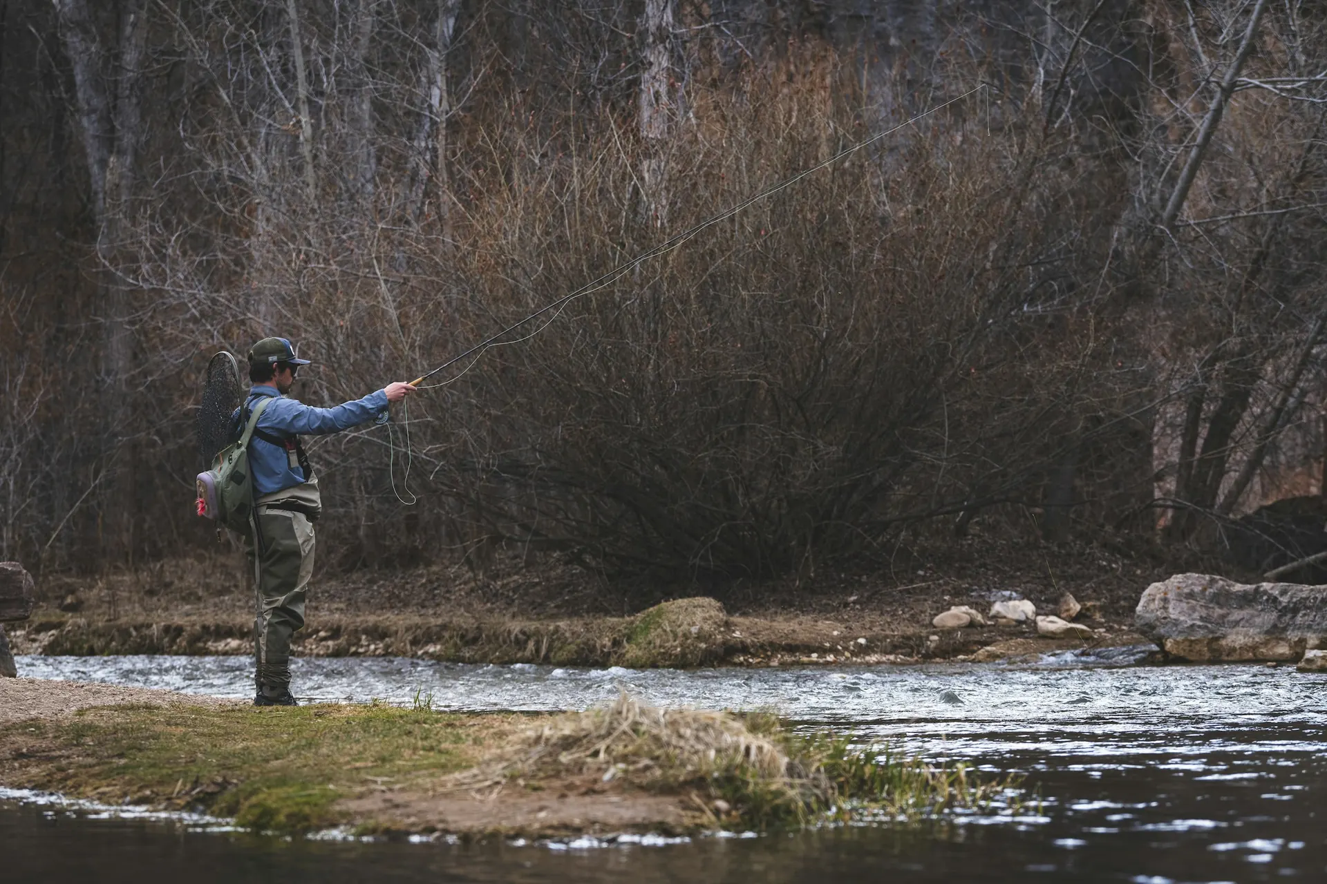 An angler fly fishing a shallow river below bare woods